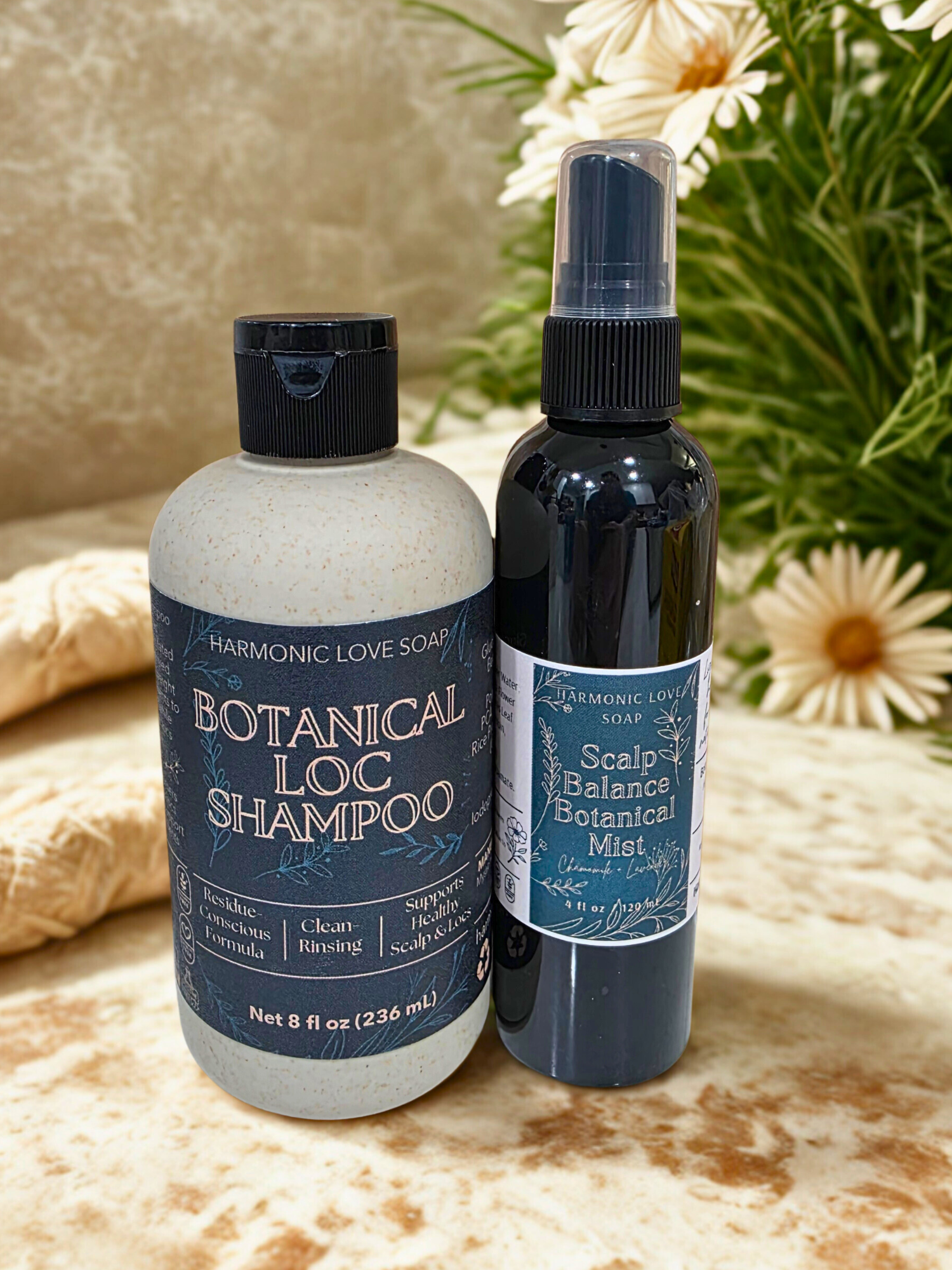 Two bottles of Harmonic Love Soap Botanical Loc Shampoo and Scalp Balance Botanical on a textured surface with flowers in the background.